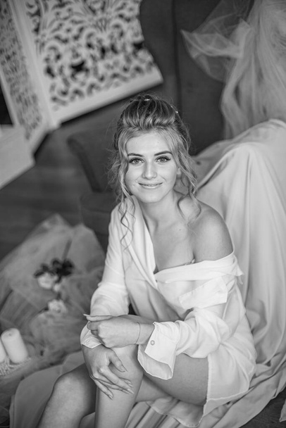 Young beautiful bride with wedding hairdo and makeup in the morning wearing a white silk negligee in the bedroom. wedding morning - 写真・画像