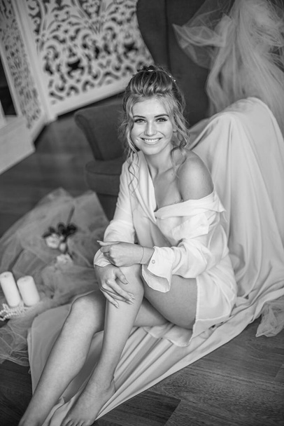 Young beautiful bride with wedding hairdo and makeup in the morning wearing a white silk negligee in the bedroom. wedding morning - Φωτογραφία, εικόνα