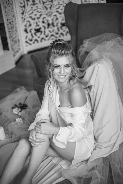 Young beautiful bride with wedding hairdo and makeup in the morning wearing a white silk negligee in the bedroom. wedding morning - Foto, imagen