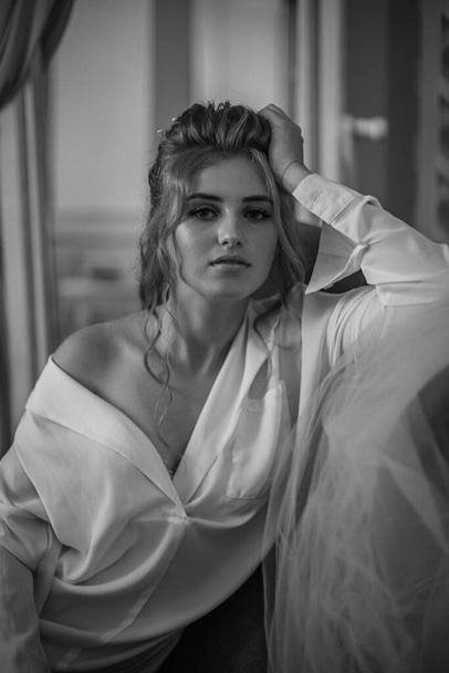 Young beautiful bride with wedding hairdo and makeup in the morning wearing a white silk negligee in the bedroom. wedding morning - Fotoğraf, Görsel