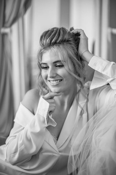 Young beautiful bride with wedding hairdo and makeup in the morning wearing a white silk negligee in the bedroom. wedding morning - Photo, image