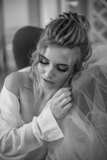 Young beautiful bride with wedding hairdo and makeup in the morning wearing a white silk negligee in the bedroom. wedding morning - Zdjęcie, obraz