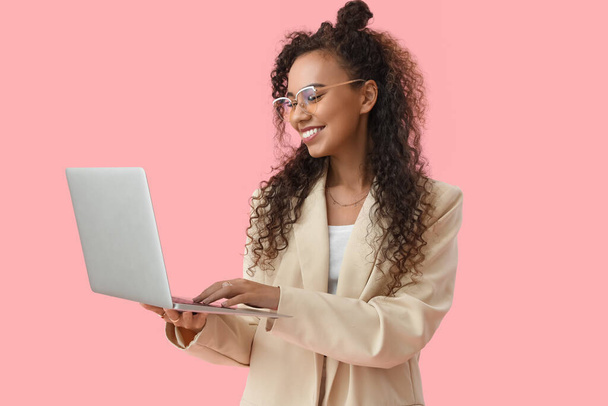 Νεαρή Αφροαμερικανή γυναίκα που χρησιμοποιεί φορητό υπολογιστή σε ροζ φόντο - Φωτογραφία, εικόνα