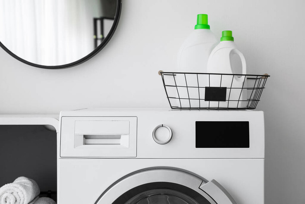 Korb mit Waschmittelflaschen auf Waschmaschine nahe Lichtwand - Foto, Bild