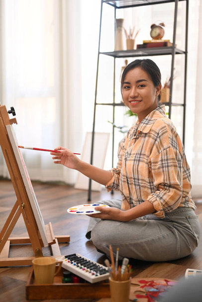 Portret artystki trzymającej paletę i obraz malarski na płótnie z farbami olejnymi w przytulnym domowym studio. - Zdjęcie, obraz