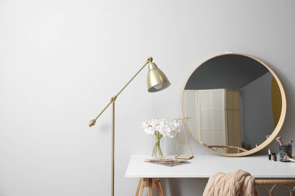 Mesa de vestir com espelho, flores em vaso, cosméticos, jóias e lâmpada perto da parede de luz - Foto, Imagem