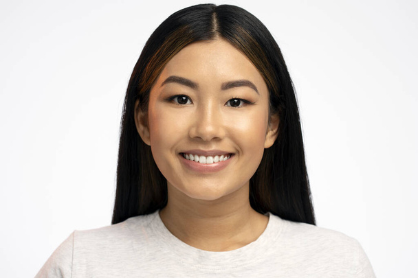 Porträt eines lächelnden ruhigen schönen asiatischen Mädchens mit langen brünetten Haaren, das mit glücklichem Gesichtsausdruck in die Kamera blickt. Indoor-Studio isoliert auf weißem Hintergrund aufgenommen  - Foto, Bild