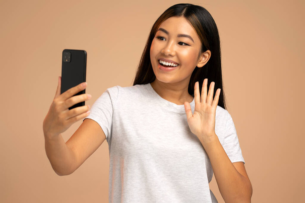 Радостная азиатская молодая женщина, использующая смартфон для видеозвонков, изолирована в коричневом, видео чате, разговаривает по мобильному телефону через видео и улыбается. Концепция видеозвонка  - Фото, изображение