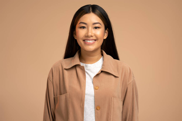Porträt eines lächelnden ruhigen schönen asiatischen Mädchens mit langen brünetten Haaren, das mit glücklichem Gesichtsausdruck in die Kamera blickt. Indoor-Studio isoliert auf braunem Hintergrund  - Foto, Bild