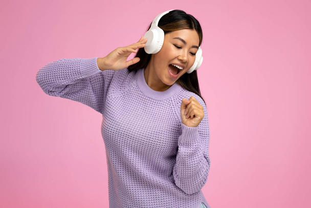 Azjatka w słuchawkach słuchająca muzyki na różowym tle. Overjoyed dziewczyna w fioletowy skoczek korzystających z ulubionych piosenek  - Zdjęcie, obraz