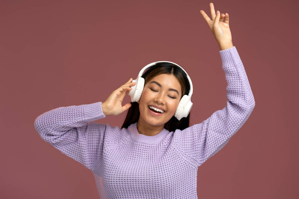 Crazy asian woman in headphones singing rejoicing and showing peace gesture isolated. Overjoyed female in violet jumper laughing and singing  - Фото, изображение
