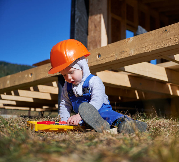 Niño pequeño jugando como constructor en el sitio de construcción. Carpintero infantil en casco naranja y mono azul aprendiendo a construir una casa de marco de madera al aire libre en un día soleado. Concepto de carpintería y taller. - Foto, imagen