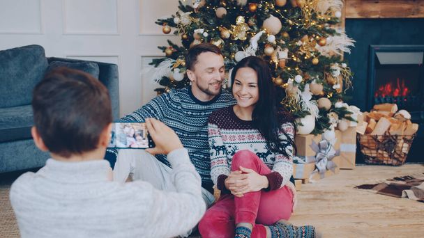 Man and woman are posing near Christmas tree while boy taking pictures with smartphone using modern device at home. People and holidays concept. - Фото, зображення