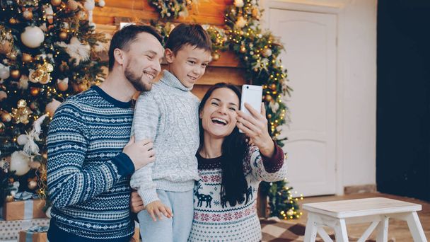Радісні батьки та дитина обговорюють онлайн-дзвінок у новорічний день за допомогою смартфона, люди носять теплий різдвяний одяг.. - Фото, зображення