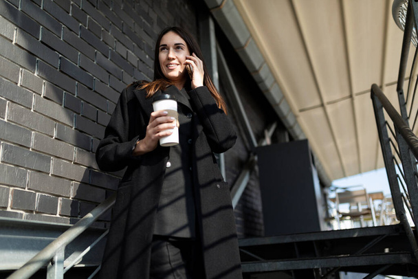 πορτρέτο μιας επιτυχημένης σύγχρονης ανεξάρτητης γυναίκας με επαγγελματικά ρούχα που μιλάει σε ένα κινητό τηλέφωνο σε μια μαύρη σκάλα με ένα φλιτζάνι καφέ στα χέρια της. - Φωτογραφία, εικόνα