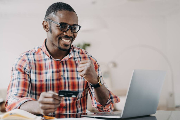 クレジットカードを持っているアフリカ系アメリカ人の男性は、ノートパソコンでオンラインバンキングサービスを使用し、はいジェスチャーを行います。現代の黒人の男は成功した送金で満足して、現金に戻る。電子銀行、電子商取引. - 写真・画像