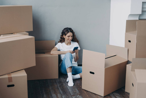 Happy young european woman with smartphone is packing boxes. Girl is chatting on mobile phone and sitting on the floor. Easy moving and shipping service order concept. - Photo, Image