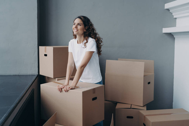 Hopeful woman is packing cardboard box. Happy hispanic girl purchasing real estate. Moving and relocation to new home. Independence, changes and new life concept. - Photo, Image