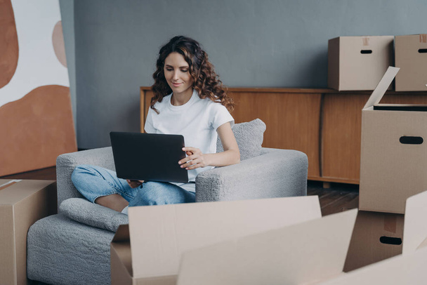 Happy european lady is sitting in armchair and working with pc. Delivery service ordering through internet. Mover among cardboard boxes is using laptop and smiling. Easy move concept. - Photo, Image