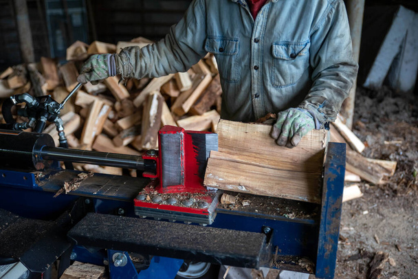 Робочий чоловік розщеплює деревину з гідравлічним розділювачем колод. Видно, що лезо ріжеться в деревину. Дефолтована дерев'яна купа на задньому плані
 - Фото, зображення