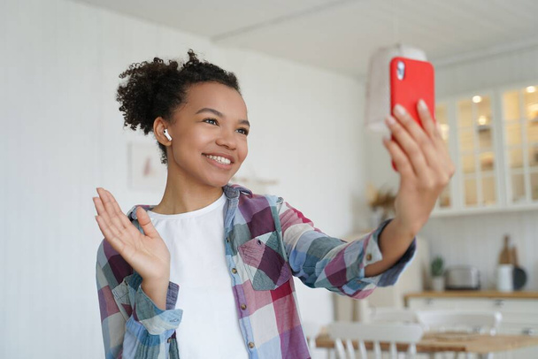 Happy biracial teen girl makes greeting hello gesture chatting online by video call, holding smartphone. Modern young mixed race lady blogger welcomes followers in social network, talking live at home - Foto, Imagen