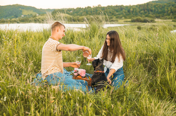 young happy couple having picnic with their french bulldog outdoors in the countryside during beautiful sunset. Romantic date near the lake - Фото, изображение