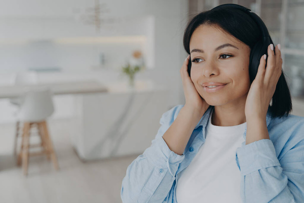 Young mixed race romantic lady is listening to radio in headphones, dreaming and smiling. Happy european girl taking pleasure from music. Enjoying weekend morning at home. Emotions and relaxation. - Фото, изображение