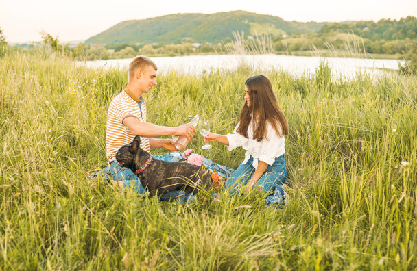 young happy couple having picnic with their french bulldog outdoors in the countryside during beautiful sunset. Romantic date near the lake - Foto, afbeelding