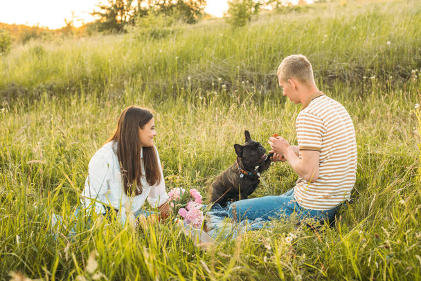 young happy couple having picnic with their french bulldog outdoors in the countryside during beautiful sunset. Romantic date near the lake - Foto, imagen