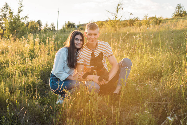 young happy couple having picnic with their french bulldog outdoors in the countryside during beautiful sunset. Romantic date near the lake - Fotó, kép