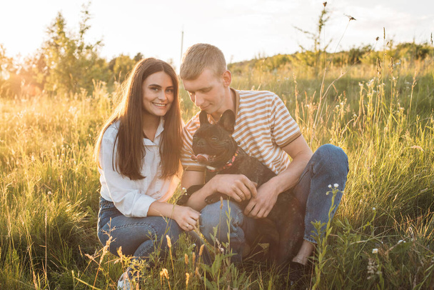 young happy couple having picnic with their french bulldog outdoors in the countryside during beautiful sunset. Romantic date near the lake - Foto, immagini
