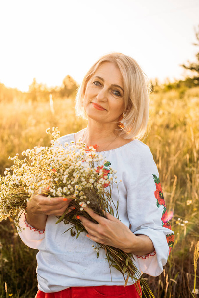 portrait of happy joyfull mature woman with a bouquet of wildflowers in countryside during beautiful sunset - Photo, image