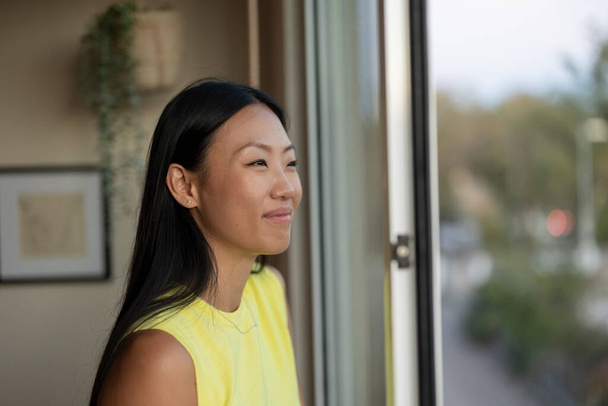 bella asiatico donna indossare un giallo camicia stand vicino il finestra felicemente guardando fuori con un sorriso su il suo faccia - Foto, immagini