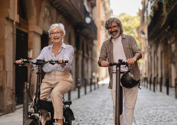 Šťastný senioři pár s zábavnou jízdu na kole a elektrický skútr přes město - Fotografie, Obrázek