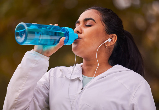 Fitness, runner and woman with bottle drinking water while running in forest for exercise, cardio workout or sports. Marathon training, thirsty or young girl with liquid hydration for wellness health. - Photo, Image