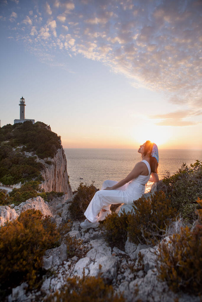 ギリシャ海の上のレフカダ島灯台日没の崖の景色に座っている幸せな女性 - 写真・画像