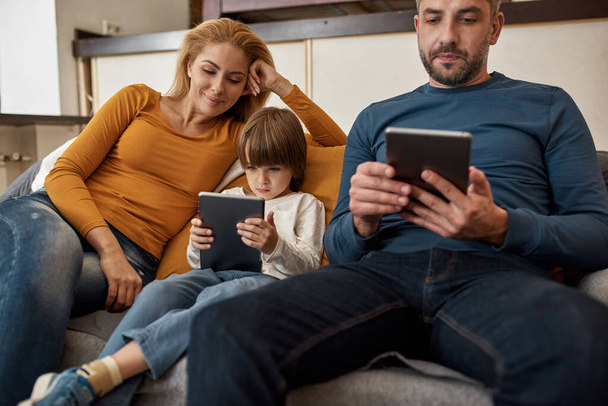 Νεαρή ευρωπαϊκή οικογένεια παρακολουθεί ψηφιακές ταμπλέτες στον καναπέ στο σπίτι. Γονείς. Η σχέση μητέρας, πατέρα και γιου και το να περνάμε χρόνο μαζί. Οικιακός ελεύθερος χρόνος και διασκέδαση. Μοντέρνο εσωτερικό διαμέρισμα - Φωτογραφία, εικόνα