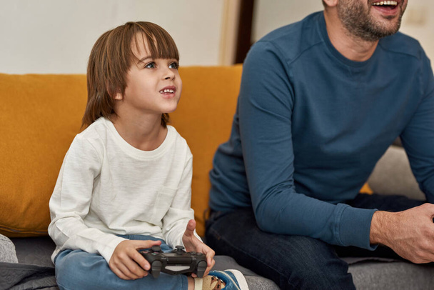Caucasian little boy and partial father playing video game with joysticks on sofa at home. Concentrated kid and man spending time together. Fatherhood and parenting. Domestic leisure and entertainment - Фото, изображение