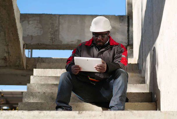 Moe Afro-Amerikaanse werknemer zit op de trap op de bouwplaats en spreekt via tablet computer via video link - Foto, afbeelding