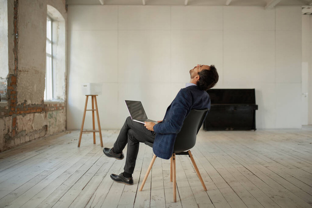 Portrait of young businessman sitting in empty room with laptop and looking at ceiling. Business failure, professional difficulties. Concept of business, challenges, work, occupation - Photo, Image