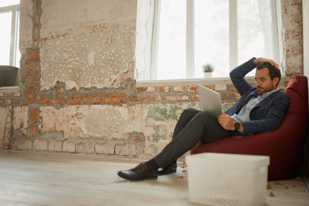 Retrato de un joven hombre de negocios con una expresión reflexiva sentado en la almohada del piso con el ordenador portátil y el trabajo. Cierre de negocio. En busca de vacantes. Concepto de negocio, desafíos, trabajo, ocupación - Foto, Imagen