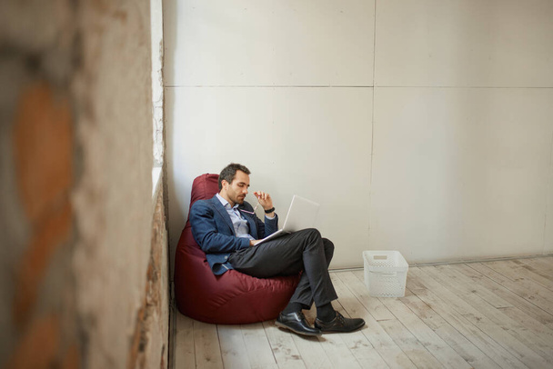 Retrato de jovem empresário com expressão pensativa sentado no travesseiro do chão com laptop e trabalhando em sala vazia. Fechamento de negócios. Conceito de negócio, desafios, trabalho, ocupação - Foto, Imagem