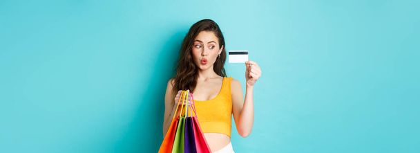 Image of happy woman shopaholic showing her plastic credit card, holding shopping bags, wearing summer clothes, standing against blue background. - Photo, Image