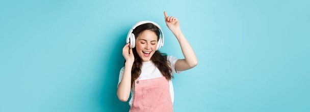Stylish millennial girl dancing in spring outfit, listening music in headphones and enjoying sound, standing over blue background. - Фото, изображение