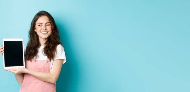 Εικόνα της ευτυχισμένης νεαρής κοπέλας που δείχνει ψηφιακή οθόνη tablet και χαμογελώντας υπερήφανοι, δείχνοντας το λογότυπό σας στην οθόνη, στέκεται πάνω από το μπλε φόντο. - Φωτογραφία, εικόνα