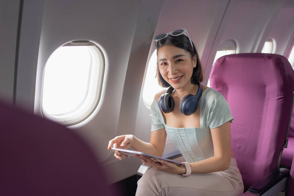 Χαρούμενη Ασιάτισσα κάθεται στο αεροπλάνο και χρησιμοποιεί τάμπλετ ενώ ταξιδεύει.. - Φωτογραφία, εικόνα