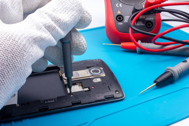 Mobile phone repair. The service technician disassembles the phone on the esd mat with a screwdriver, multimeter in the background - Foto, afbeelding