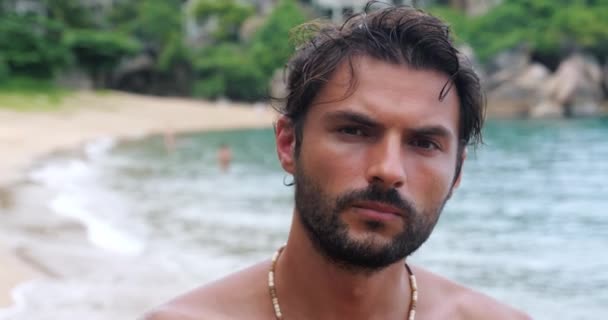 Confiado chico caucásico con el torso desnudo, mirando con una mirada perspicaz, disfrutando tomando el sol en la playa tropical - Metraje, vídeo