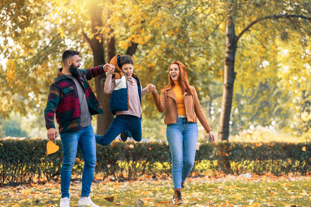 Jóvenes padres felices divirtiéndose con su hijo mientras corren en el parque durante el día de otoño. - Foto, imagen