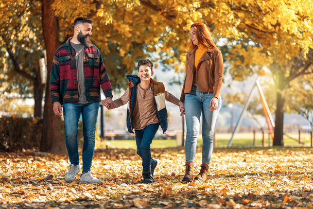 秋の日に公園を走りながら楽しい時間を過ごしている若い幸せな両親. - 写真・画像